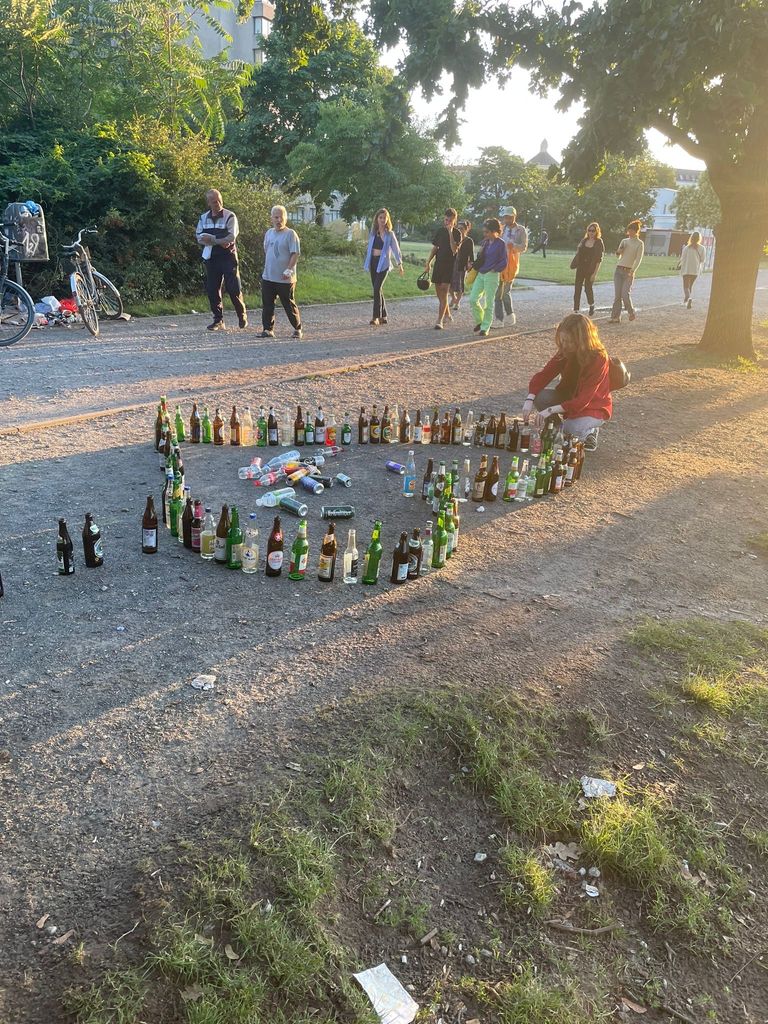 Vocal performance student Gwyneth Rix seen assembling glass bottles into a heart shape on an off road in Germany. In the background, people walk by observing Gwyneth's art