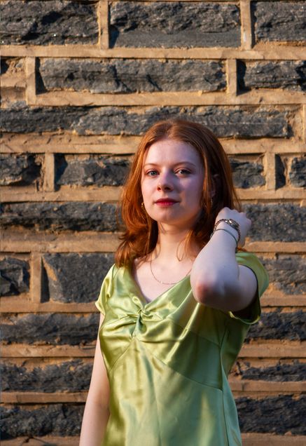 Vocal performance undergraduate student Gwyneth Rix, standing in front of a brick wall. She is wearing a light green satin top and looking at the camera with a slight smile.