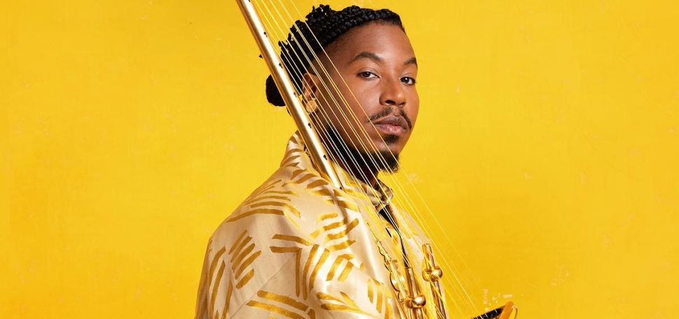 A person wearing a gold and cream patterned outfit poses against a bright yellow background, holding a stringed musical instrument with golden accents. The individual has braided hair styled neatly and looks confidently toward the camera, creating a vibrant and artistic composition.