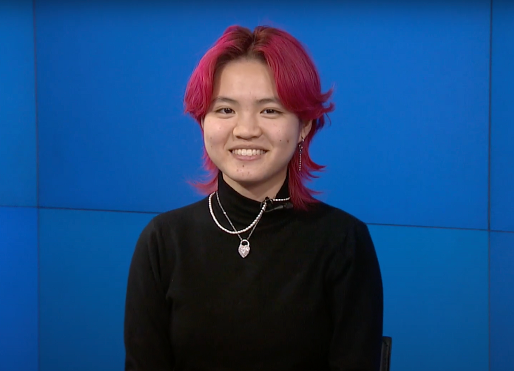 BU College of Fine Arts BA music major Miya Menscher with vibrant pink, layered hair sits in front of a bright blue background, smiling warmly at the camera. They are wearing a black turtleneck sweater, accessorized with layered silver necklaces, one featuring a heart-shaped pendant. They also wear dangling earrings, adding to their stylish look.