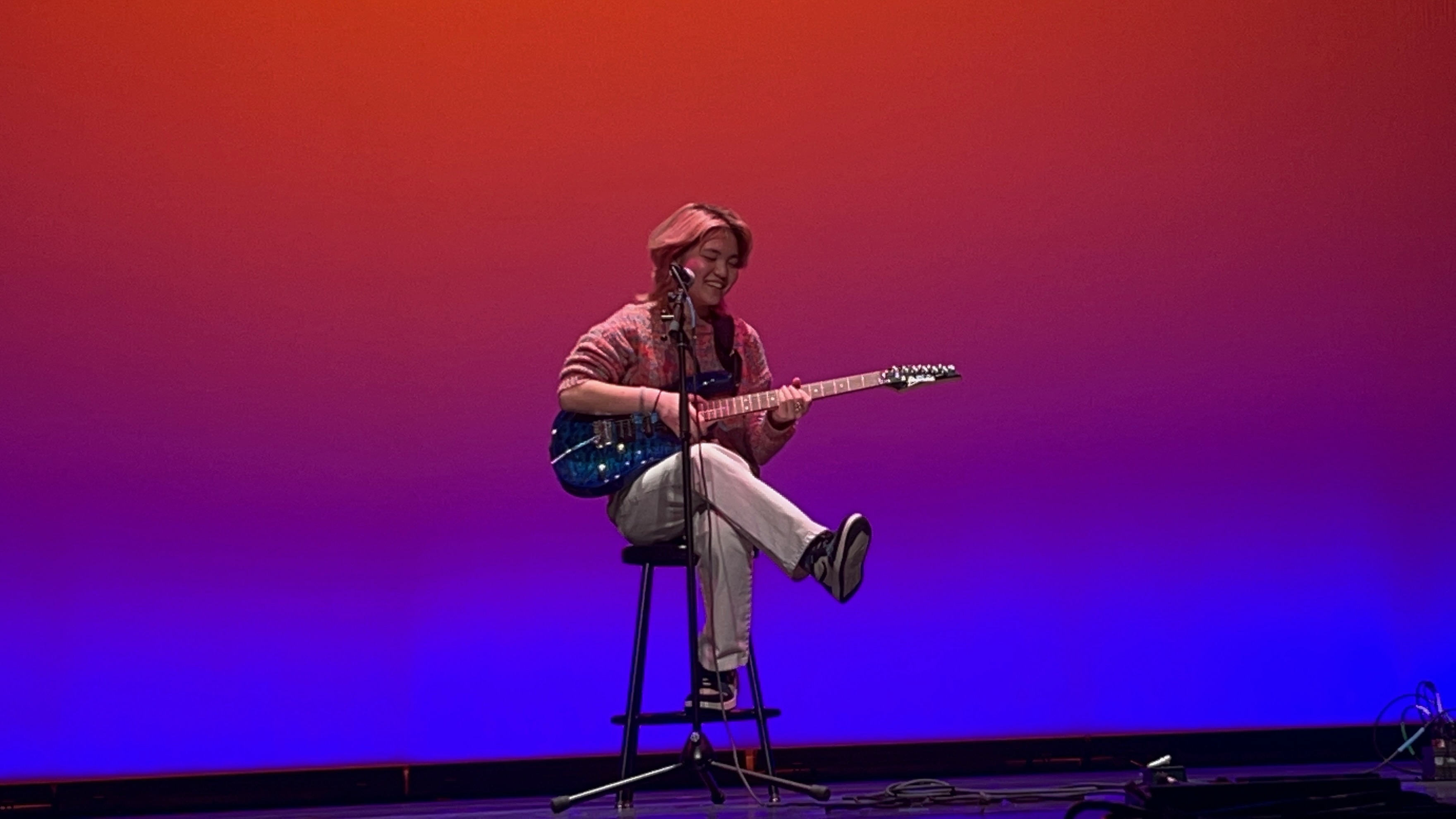 Miya Menscher with short, wavy hair sits on a stool onstage, playing a blue electric guitar and singing into a microphone. They are wearing a cozy, patterned sweater and white pants, with one foot playfully resting on the stool’s rung. The stage lighting creates a gradient backdrop, transitioning from deep purple at the bottom to warm orange at the top, adding a dramatic and artistic effect to the scene.