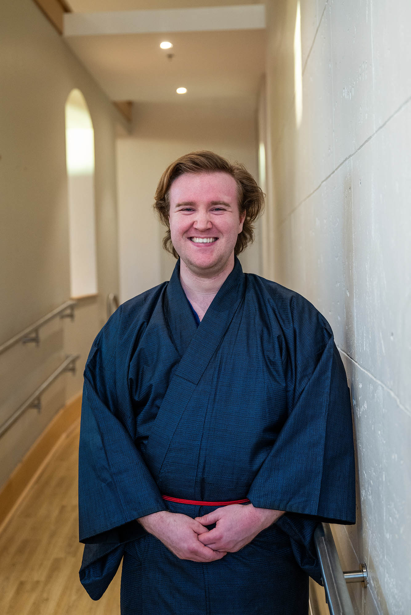 Christopher Ellars (CAS’24, CFA’25), a dual Japanese and voice performance major with medium-length, wavy blond hair stands in a softly lit hallway, smiling warmly at the camera. He is wearing a traditional dark blue Japanese kimono with a red obi belt. The background features cream-colored walls, a wooden floor, and handrails along the sides, giving the space a serene and elegant atmosphere.