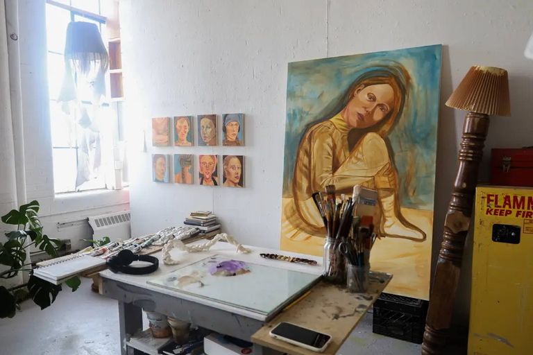 Inside the studio of Scout Curtin, a first-year painting student at Boston University. Scout’s studio has a large painting of a woman sitting with her arms wrapped around her knees, leaning to one side, placed against a white wall. To the left, a series of smaller portrait paintings are displayed in a grid. An art table covered with brushes, paint tubes, and sketchpads is positioned in the center. Sunlight streams through tall windows draped with light fabric, casting a soft glow. A leafy plant sits by the window, adding a touch of greenery. A yellow cabinet labeled "FLAMMABLE KEEP FIRE AWAY" is on the right, next to a vintage floor lamp with a tilted shade.
