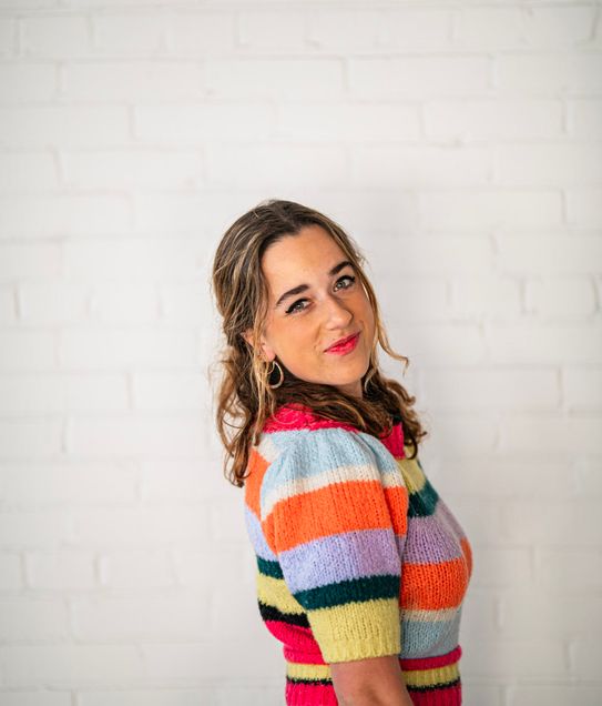 Boston University vocal performance major Sadie Habas (CFA'25) wearing a vibrant, colorful, striped knit top poses against a white brick wall. She looks over her shoulder with a playful smile and bright red lipstick. Her wavy hair cascades loosely as she leans slightly to one side. The cheerful colors of her outfit contrast beautifully with the minimalist background, creating a lively and energetic portrait.