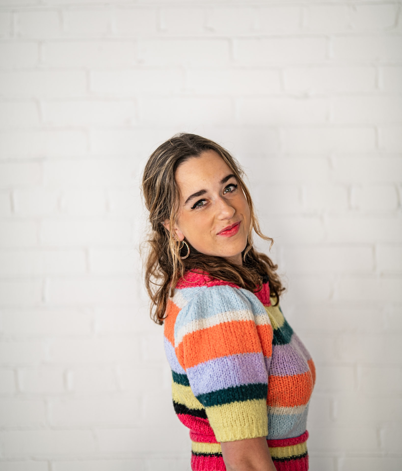 Boston University vocal performance major Sadie Habas (CFA'25) wearing a vibrant, colorful, striped knit top poses against a white brick wall. She looks over her shoulder with a playful smile and bright red lipstick. Her wavy hair cascades loosely as she leans slightly to one side. The cheerful colors of her outfit contrast beautifully with the minimalist background, creating a lively and energetic portrait.