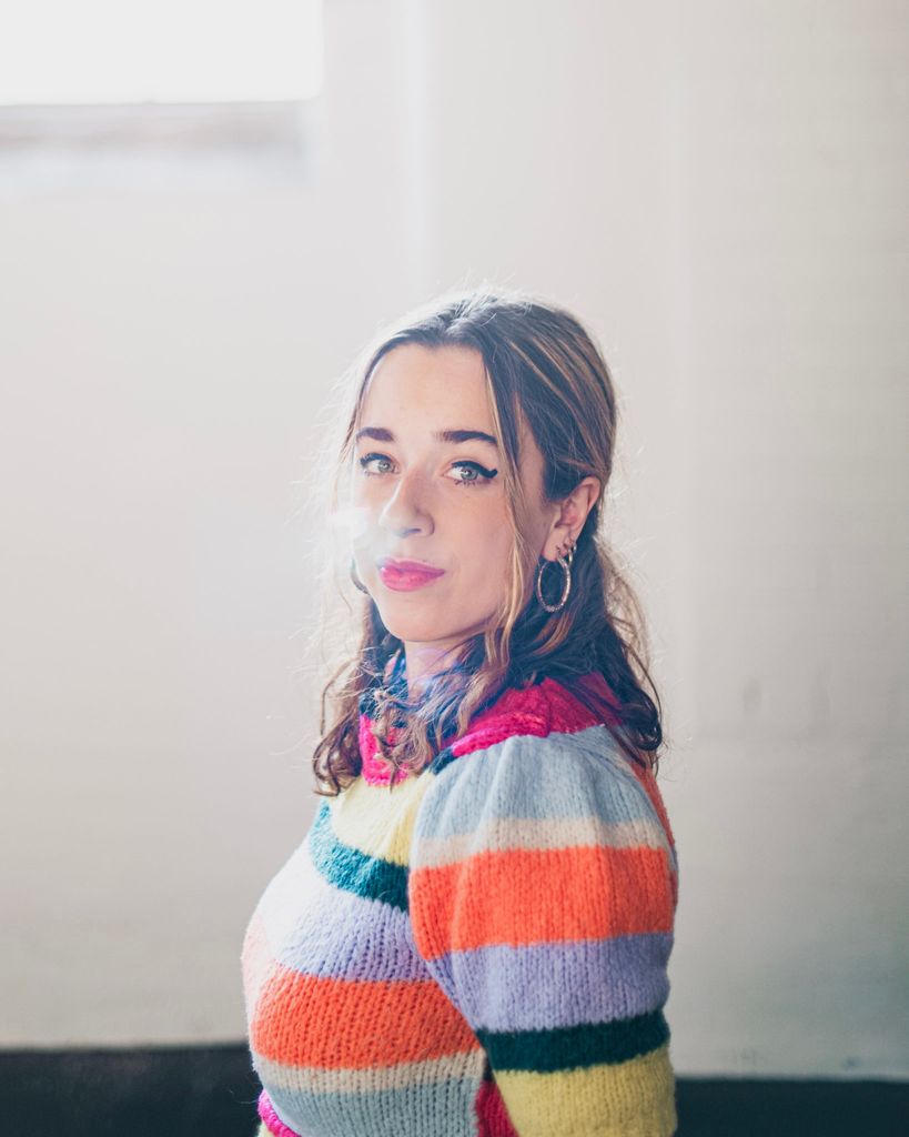 Boston University vocal performance major Sadie Habas (CFA'25) wearing a vibrant, striped knit sweater looks thoughtfully at the camera with a soft, relaxed expression. She stands in a softly lit room with sunlight streaming through a high window, creating a gentle glow around her face. Her curly hair cascades over her shoulders, and she wears hoop earrings. The serene, airy ambiance adds a contemplative mood to the portrait.