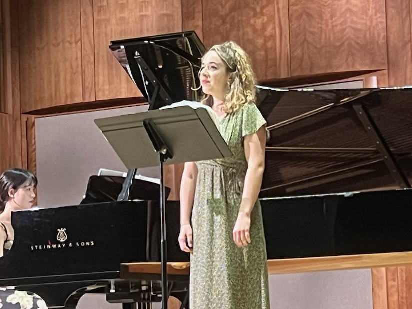 A vocalist performs on stage next to a Steinway & Sons grand piano, with a pianist accompanying in the background. The singer wears a light green floral dress and stands confidently behind a music stand, her expression engaged and emotive. The wooden paneling and warm lighting of the concert hall provide an elegant and intimate backdrop, enhancing the recital atmosphere.