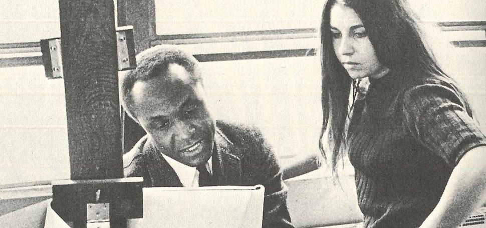 A black-and-white photo of the late artist and Boston University professor John Wilson and a student engaged in a discussion. Wilson, seated on the left, holds a large sheet of paper on an easel, pointing and speaking animatedly. The student, standing with one hand on her hip, listens attentively, her long hair falling over her shoulder. They are in a bright studio space with large windows in the background, casting soft light over the scene, emphasizing the collaborative and educational atmosphere.