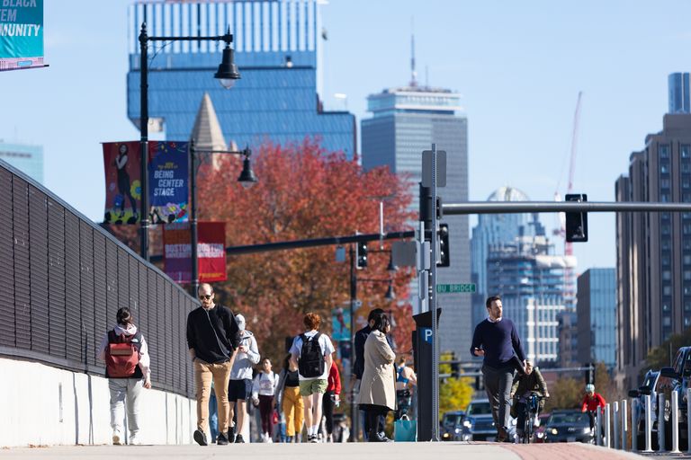 People walk along the BU Bridge on a sunny day, with the Boston skyline in the background. The Prudential Tower and other skyscrapers rise behind the autumn-colored trees. Banners for Boston University hang from lampposts, and pedestrians, some wearing backpacks and earbuds, walk along the sidewalk. Cyclists ride in the bike lane, and cars are visible in the distance.