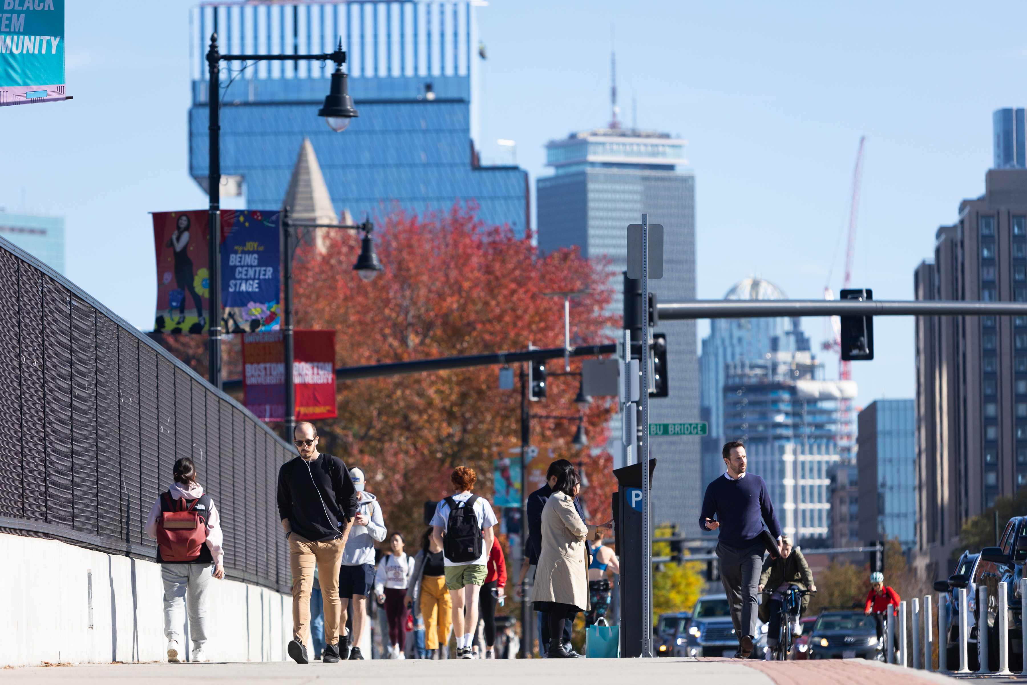 People walk along the BU Bridge on a sunny day, with the Boston skyline in the background. The Prudential Tower and other skyscrapers rise behind the autumn-colored trees. Banners for Boston University hang from lampposts, and pedestrians, some wearing backpacks and earbuds, walk along the sidewalk. Cyclists ride in the bike lane, and cars are visible in the distance.
