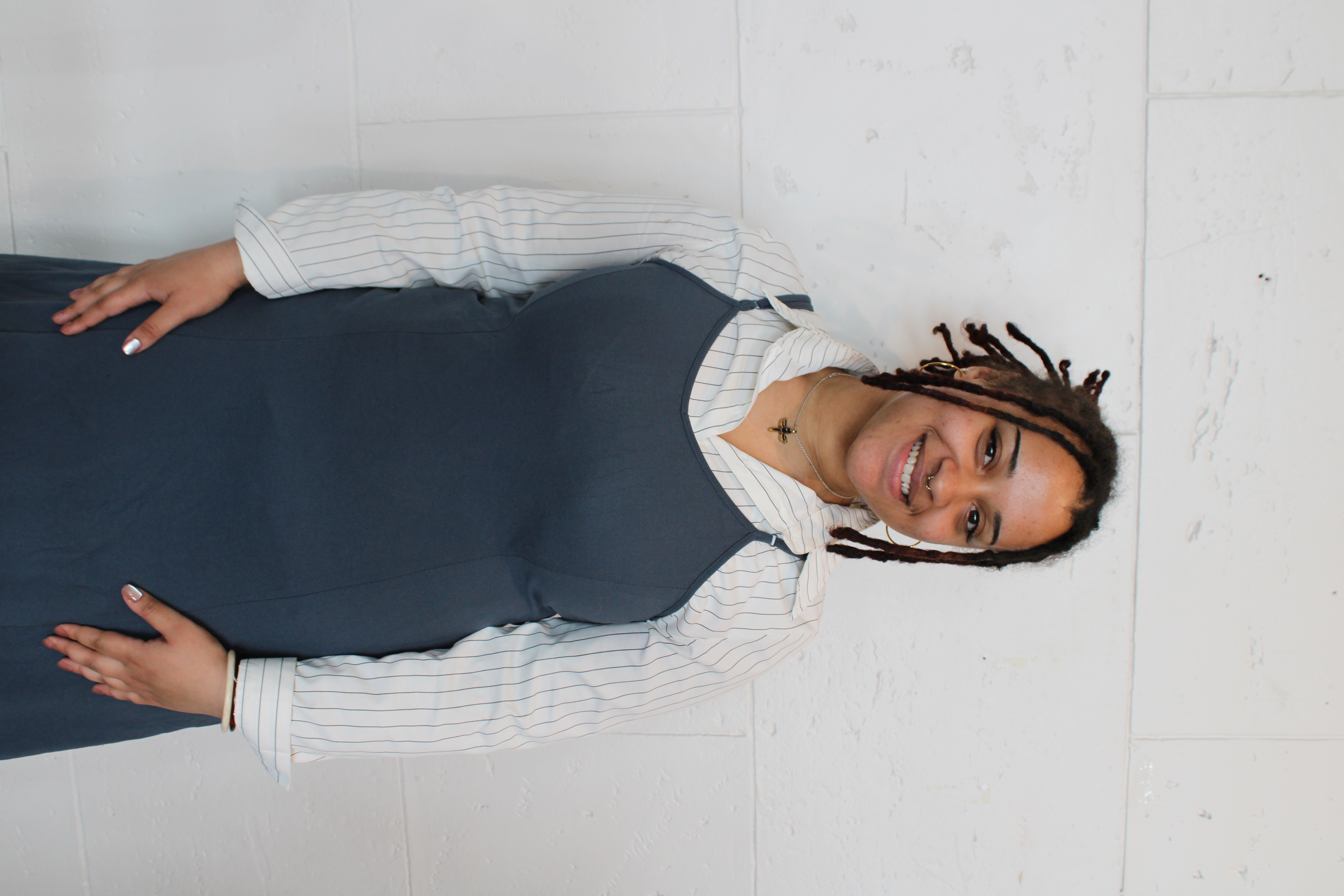 A portrait of Nyla Mawire standing against a white wall, smiling warmly at the camera. Nyla is a tuba performance undergraduate student at Boston University School of Music and the University's 2025 Kahn Career Entry Award recipient. She wears a blue dress layered over a white pinstriped shirt and has her hands loosely clasped in front of her.