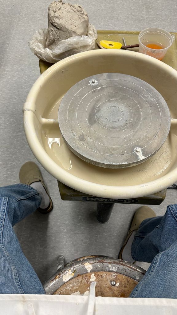 A first-person view of a seated individual preparing to work on a pottery wheel. In view are a lump of wet clay, a water container, sculpting tools, and the circular pottery wheel on a Brent base. The person is wearing jeans and slippers.