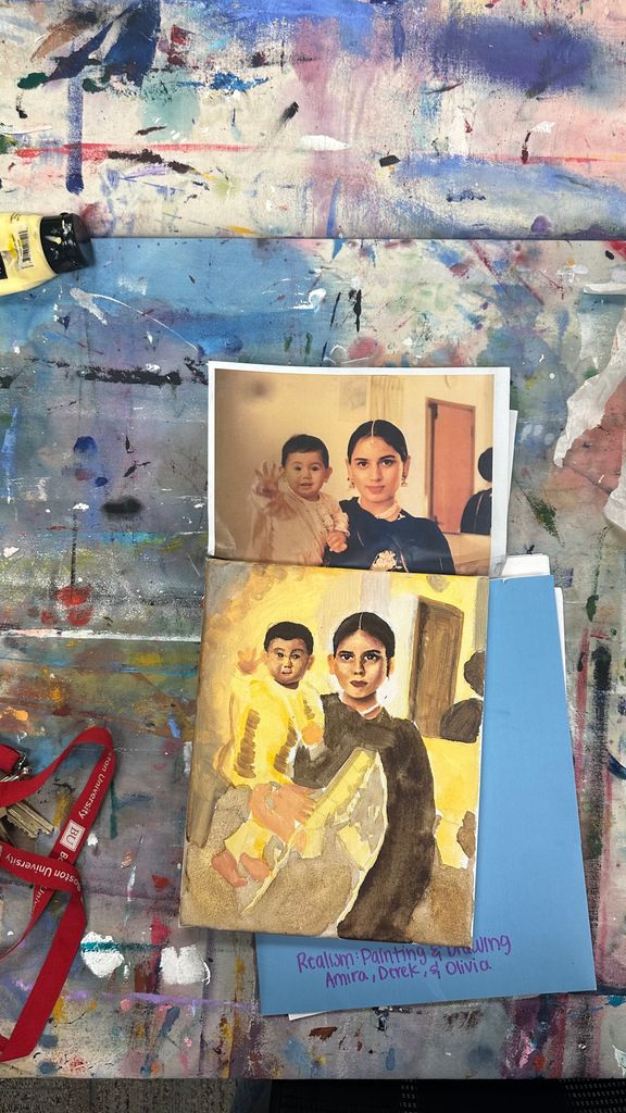 A painting in progress on a messy studio table, depicting a woman holding a child. Above it is the reference photo of the same scene. The workspace is covered in splatters of paint and includes a blue folder labeled “Realism: Painting & Drawing, Amira, Derek, & Olivia.”