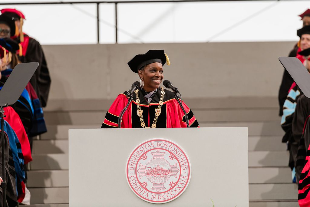 Boston University President Melissa L. Gilliam, dressed in red and black academic regalia with a gold medallion necklace, stands at a podium bearing the BU seal and smiles while delivering remarks at the 2025 Commencement ceremony. Faculty in academic dress are seated in the background.