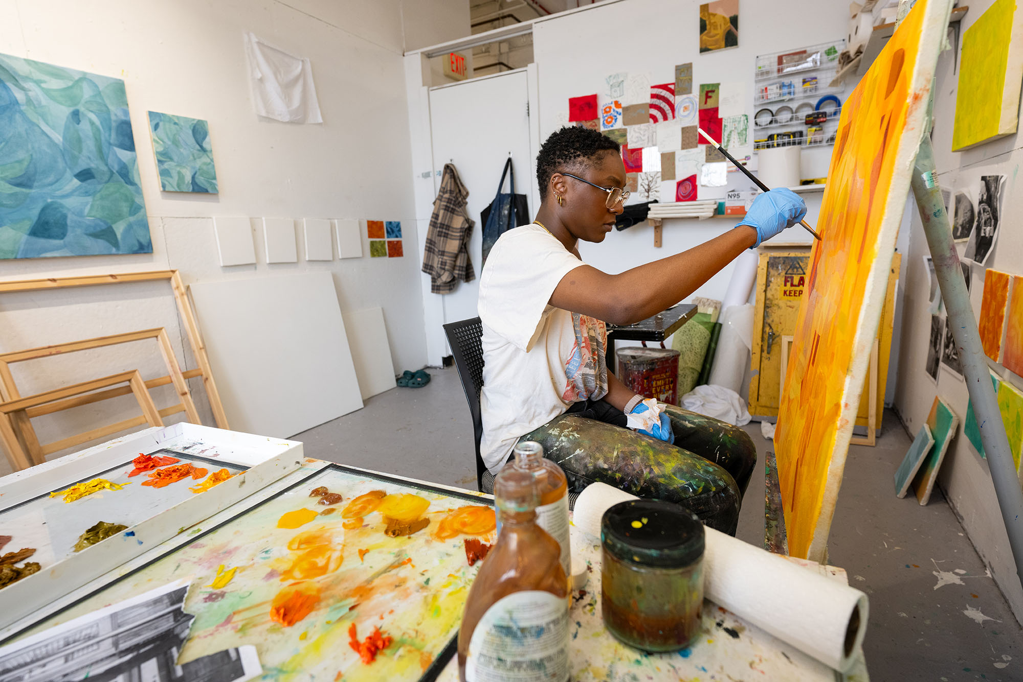 Raquel Philippe, a Boston University MFA Painting student, sits in her studio working on a large canvas in vibrant shades of orange. She wears paint-splattered pants, a casual graphic t-shirt, and blue gloves, focusing intently on her brushwork. Her studio is filled with colorful artwork, paint supplies, and blank canvases leaning against the wall.