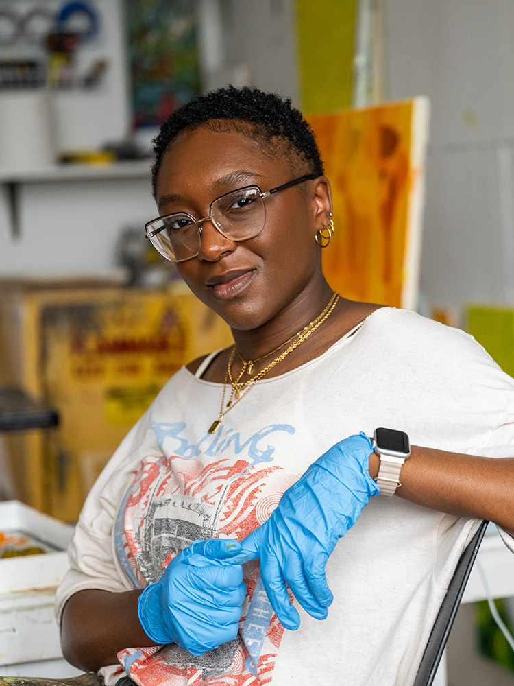 Boston University MFA Painting student Raquel Philippe sits in a studio space wearing a graphic T-shirt, glasses, and blue disposable gloves. She looks confidently at the camera, with one arm crossed and the other adjusting a glove. She wears layered gold necklaces, hoop earrings, and an Apple Watch. Behind her are colorful abstract artworks and studio materials.
