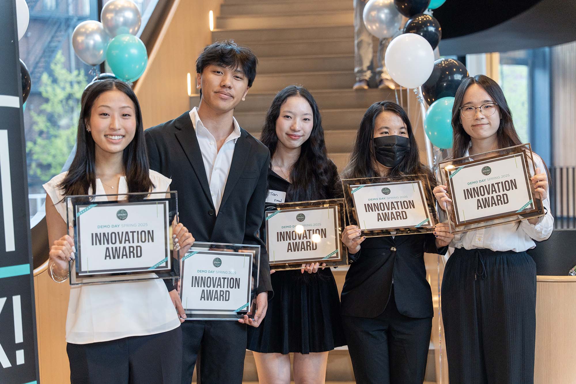 Five students stand smiling and holding framed certificates labeled “Innovation Award” at BU Spark! Demo Day Spring 2025. They are dressed in business or semi-formal attire, and balloons decorate the staircase behind them.
