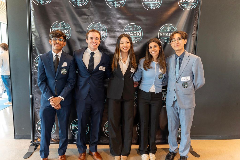 Five students stand in a row, smiling and dressed in business attire, in front of a BU Spark! branded backdrop. They wear name tags and Spark! event buttons, posing together at Demo Day Spring 2025.