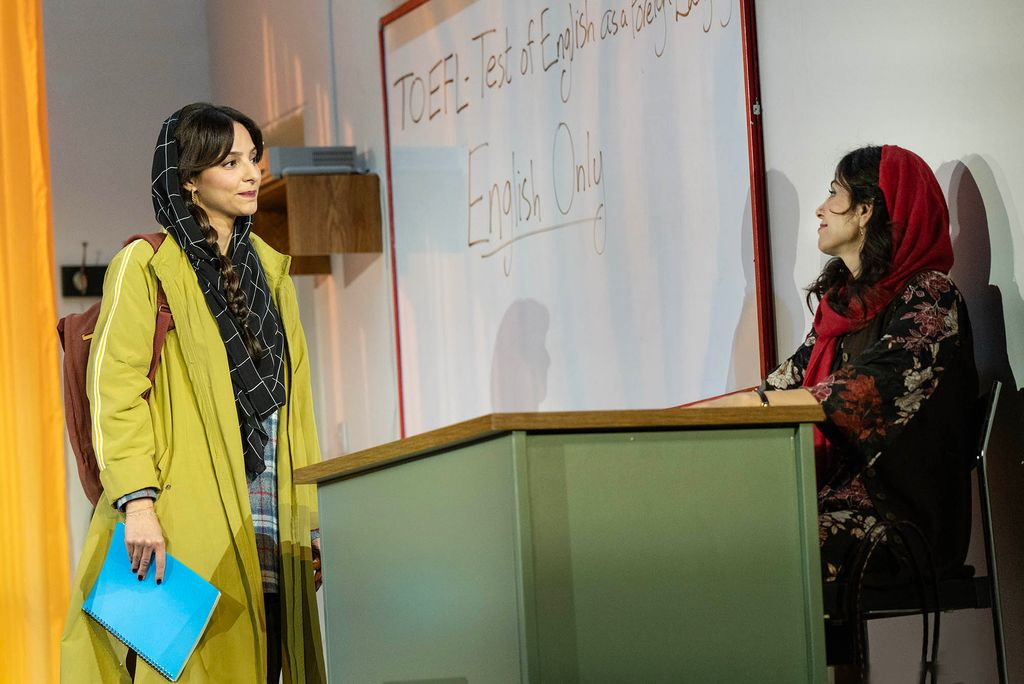 A scene from the Broadway show "English" depicting two individuals in a classroom setting. One person, dressed in a bright yellow coat and carrying a blue notebook, stands in front of a green desk while engaging with another person seated behind it. The seated individual is wearing a black floral garment and a red headscarf. Behind them is a white board with the words "TOEFL: Test of English as a Foreign Language" and "English Only" written on it, setting the context for the scene. The backdrop features warm lighting that adds to the atmosphere.