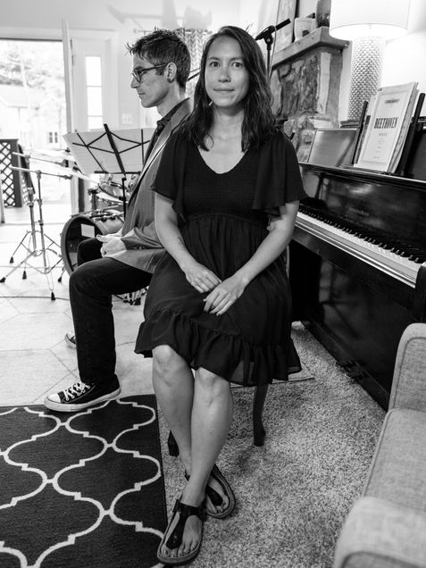 Black and white image of Gareth Dylan Smith, a Boston University College of Fine Arts School of Music assistant professor of music, music education, and pianist Austina Lee (CFA'23) sitting back to back indoors, with a piano and music stand in the background.