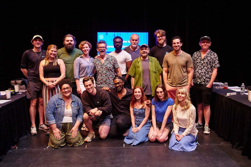 A group of seventeen people are gathered on a stage in front of a black curtain and a lit screen. Some people are standing while others are kneeling or sitting on the floor. The individuals are casually dressed in a variety of clothing styles, including shorts, t-shirts, dresses, and casual shirts. Tables with papers, cups, and bottles line the sides of the stage, suggesting a meeting or workshop setting. The atmosphere appears to be relaxed and informal.