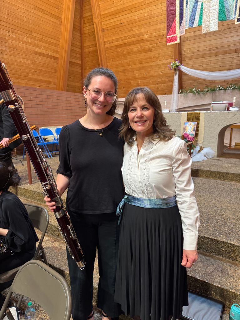 Nora Cannizaro (CFA’25), a BU bassoon performance alum, and Barb Raney, Boston University School of Music Assistant Director, Student Services, and Academic Advisor for CFA undergraduate music students, standing side by side inside a wooden-paneled room at the Immanuel Lutheran Church, the meeting place of Bristol Chorale. Nora, on the left, is wearing a black outfit and holding a large bassoon, while Barb, on the right, is dressed in a white blouse with a blue sash and a black pleated skirt. There are chairs and a small altar decorated with colorful banners and flowers in the background.