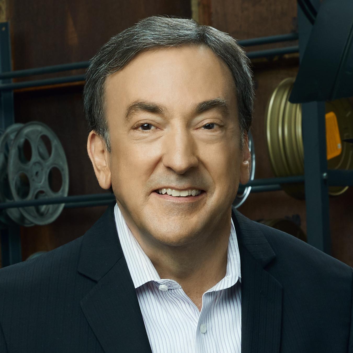 Award-winning producer Peter S. Del Vecho (CFA'80) with short gray hair, wearing a black suit jacket over a white collared shirt with light vertical stripes, stands in front of shelves holding several large reels in a dimly lit room.