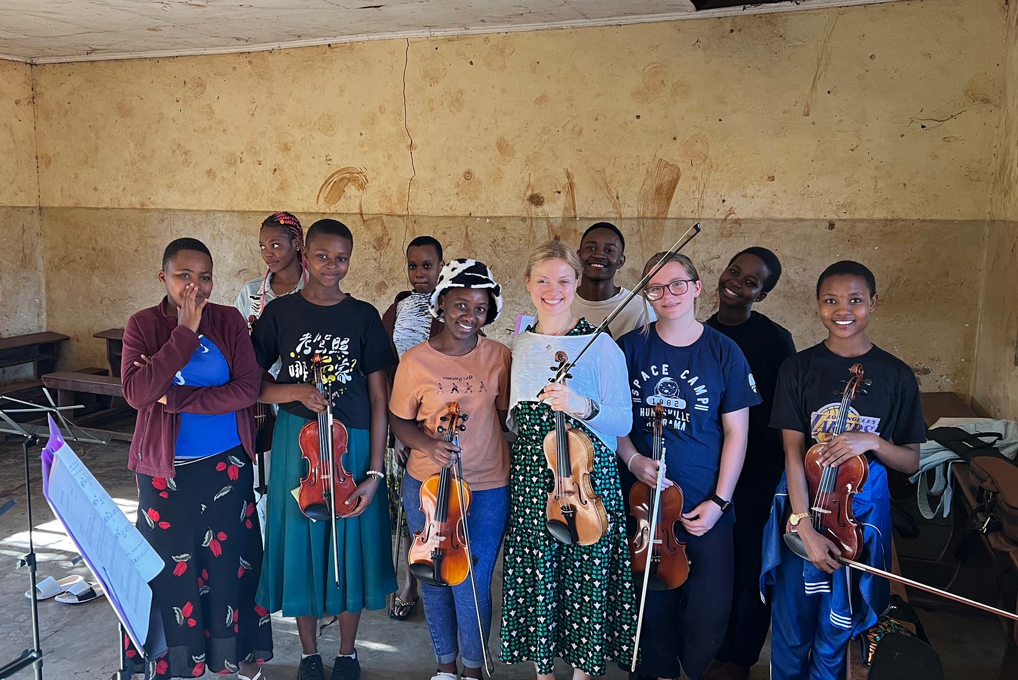 Group of young musicians in a classroom setting, some holding violins and bows, standing in a line facing the camera. There are music stands with sheet music in the foreground, and the room has worn, beige walls with visible marks and cracks. The floor appears to be concrete, and there are wooden benches against the walls. The atmosphere suggests a music practice or lesson in progress.