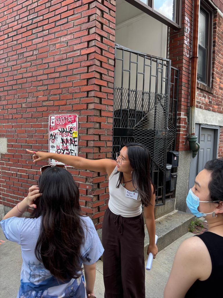 A group of three people standing by a brick wall and a gated entrance on a city sidewalk. Megan Kwan is pointing towards something while holding a rolled-up paper.