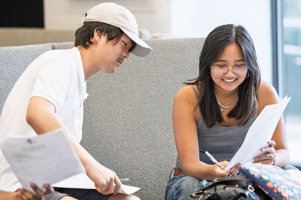 Two people sitting on a couch and looking at papers. Megan Kwan is holding a pen, while the other leans in, appearing to discuss the documents.