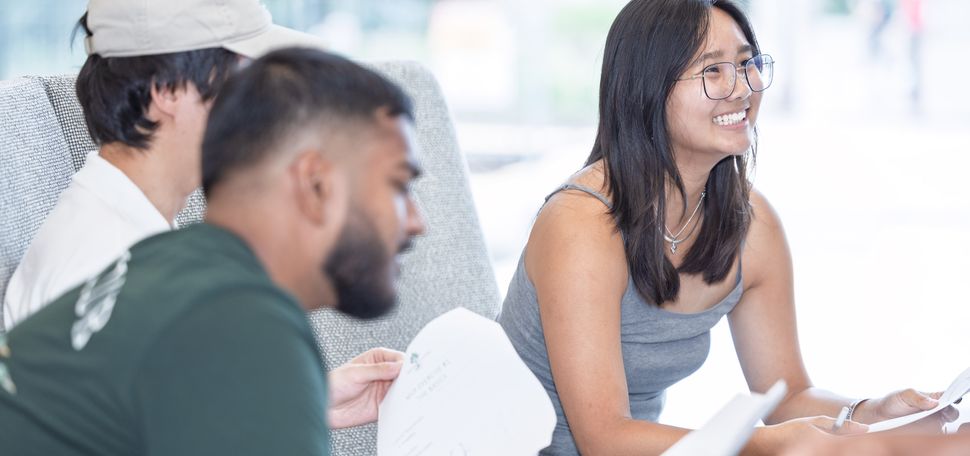 Three people sitting on a couch with papers in hand, engaged in discussion. One person is in the foreground, another person is partially visible on the side, and the third person (Megan Kwan) is seated upright with a pen.