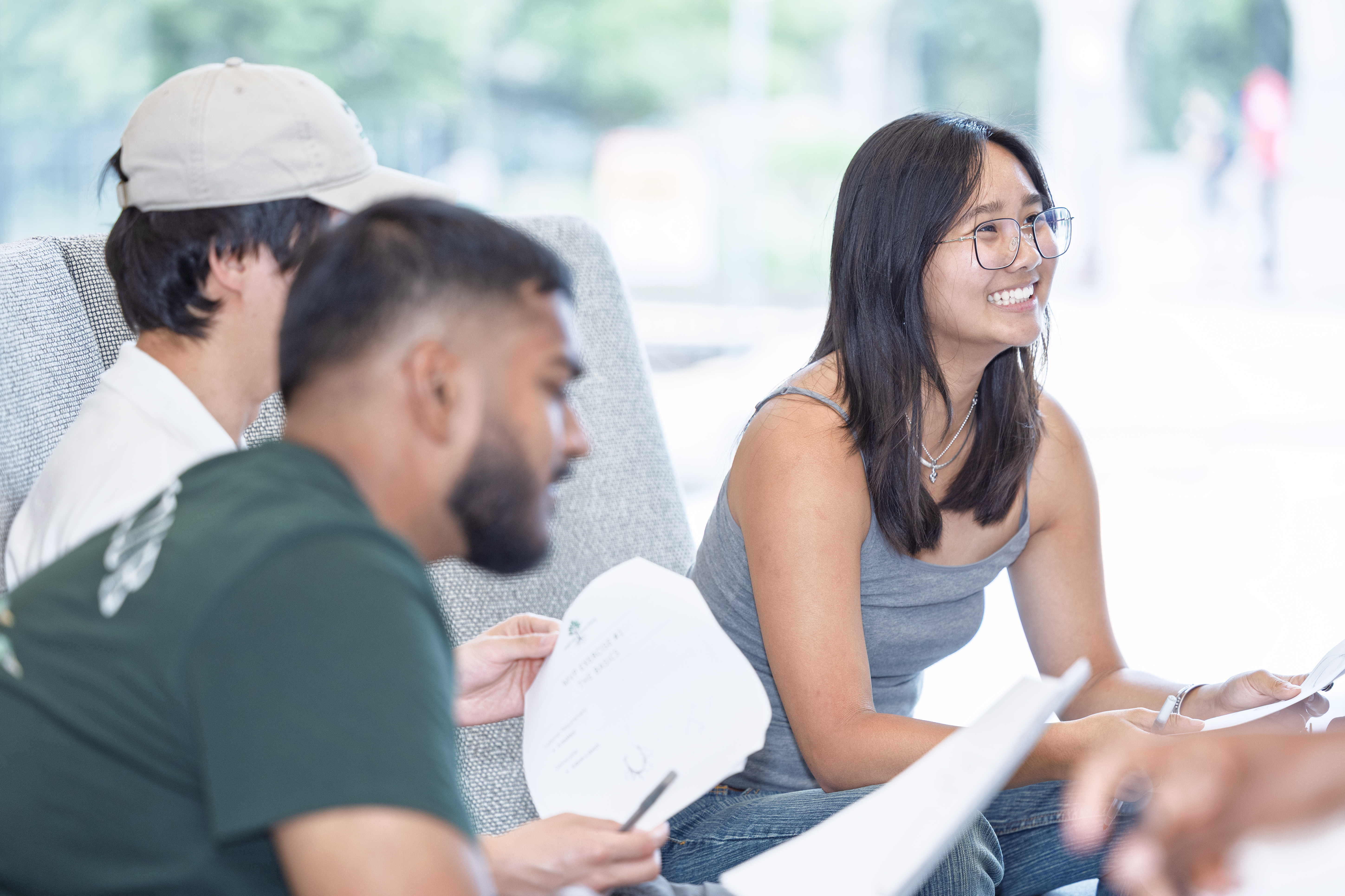 Three people sitting on a couch with papers in hand, engaged in discussion. One person is in the foreground, another person is partially visible on the side, and the third person (Megan Kwan) is seated upright with a pen.