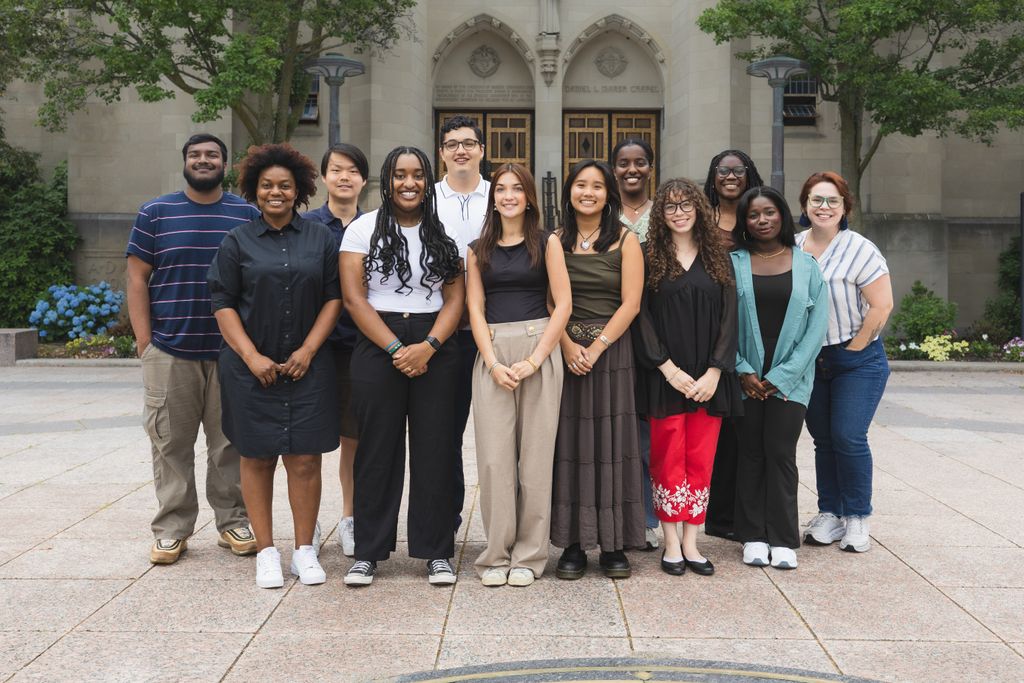 A group of twelve people standing together outside in front of a stone building at Boston University with ornate wooden doors. The group consisting of Innovate@BU Enlight Summer Fellows is posing for a photo on a paved area with greenery and flowers around.