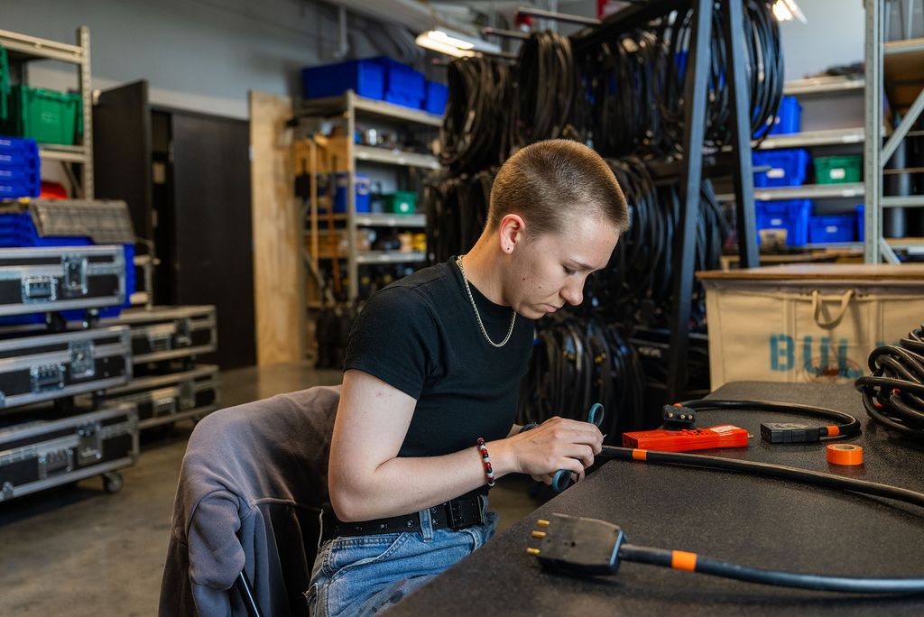 A person with short hair wearing a black t-shirt and jeans is seated at a workbench in a workshop, working on a cable with tools and electrical tape. Shelves filled with coiled cables, blue and green storage bins, and equipment cases are visible in the background. The person appears focused on their task in an industrial-type setting.