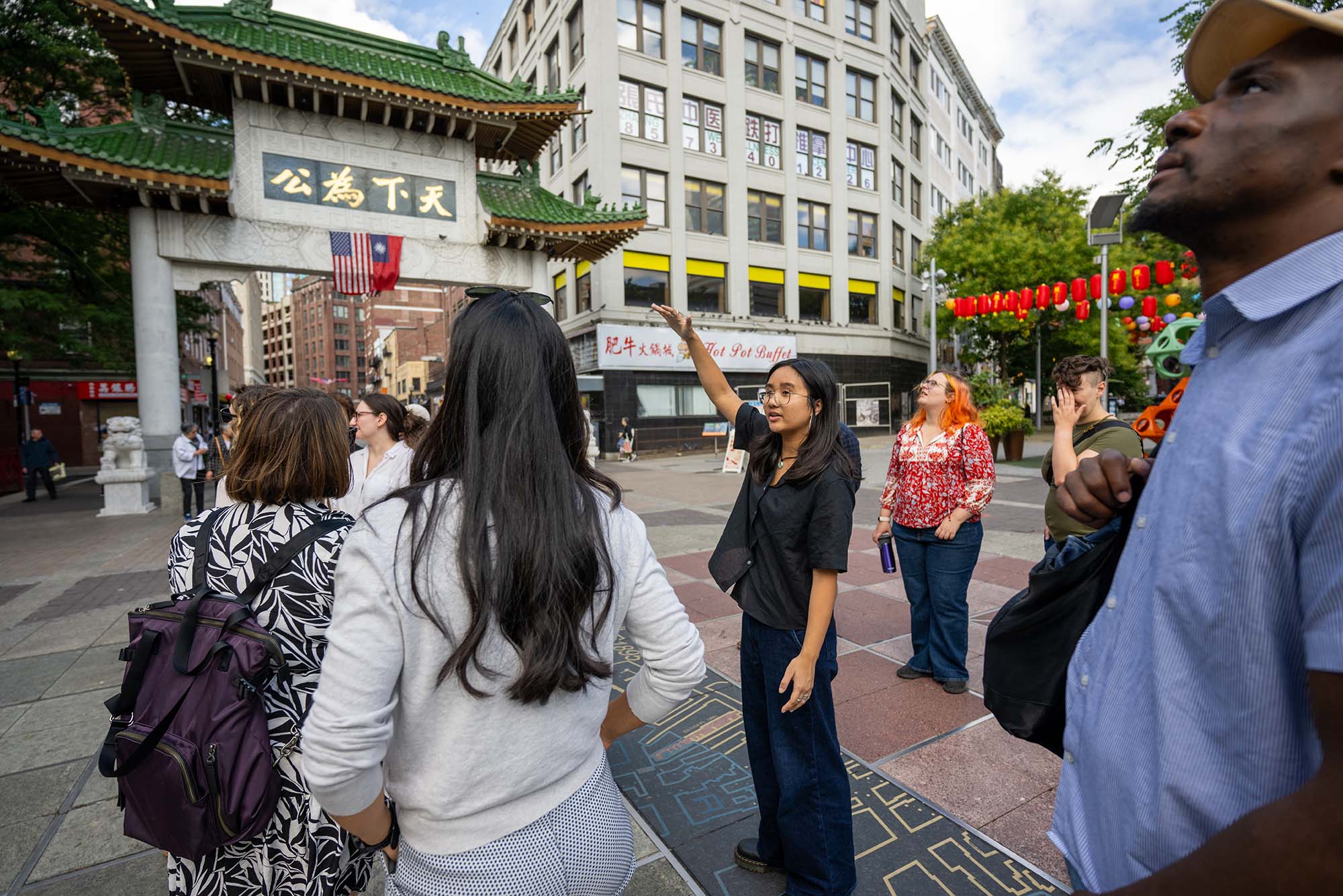 A group of people stand outdoors in an urban area near a traditional Chinese-style gate with green tiled roof and Chinese characters. The group appears to be engaged in a conversation or tour, with one woman in a black shirt gesturing and speaking. The surrounding area includes buildings with multiple windows and a "Hot Pot Buffet" sign. Red lanterns hang in the background beside some trees. One person carries a purple backpack, and the weather looks clear with blue sky and some clouds.