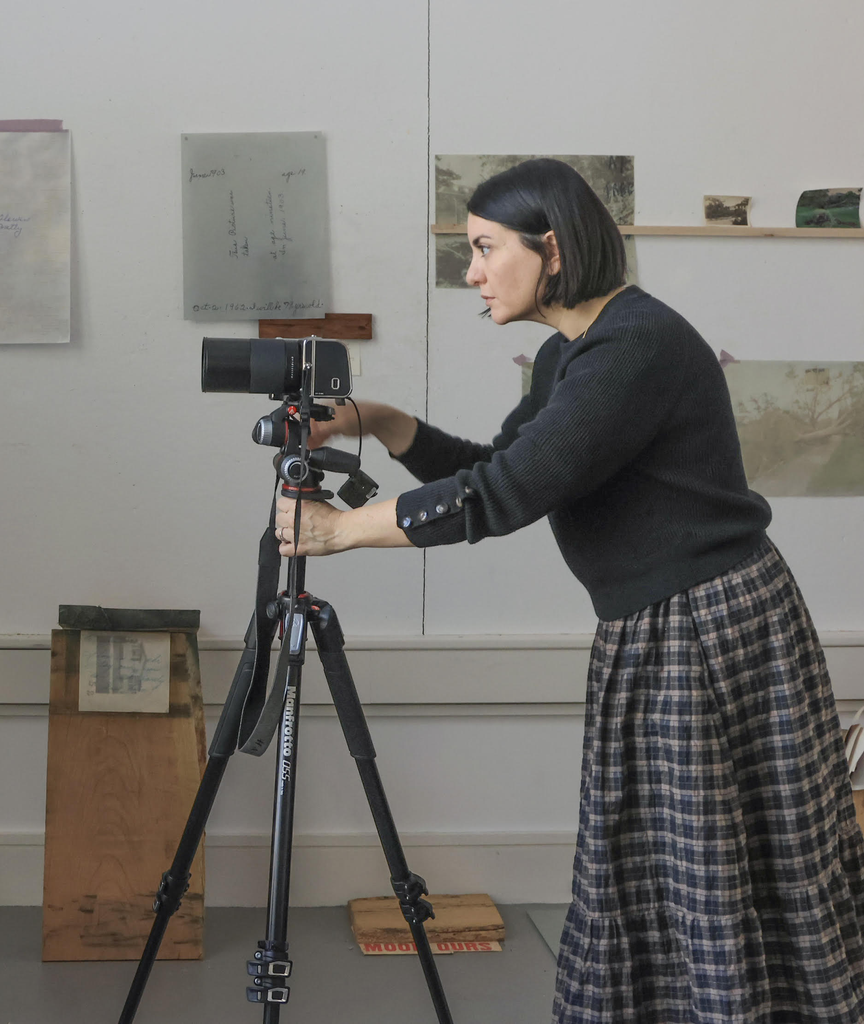 Toni Pepe, Chair, Department of Photography and Associate Professor of Art at Boston University College of Fine Arts School of Visual Arts, is wearing a black sweater and a plaid skirt while adjusting a camera mounted on a tripod in a room with several papers and pictures taped to the wall in the background.