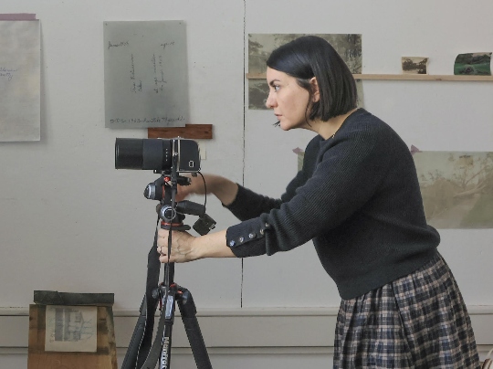 Toni Pepe, Chair, Department of Photography and Associate Professor of Art at Boston University College of Fine Arts School of Visual Arts, is wearing a black sweater and a plaid skirt while adjusting a camera mounted on a tripod in a room with several papers and pictures taped to the wall in the background.