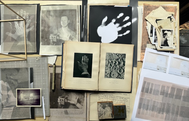 A collection of vintage photographs, printed images, and books spread out on a flat surface. The central open book features black and white images, one of a hand and the other of a textured surface. Surrounding materials include a photo of a handprint, handwritten notes, and various other printed and photographic items arranged in a collage-like manner. A wooden frame and some tools are also visible among the items.