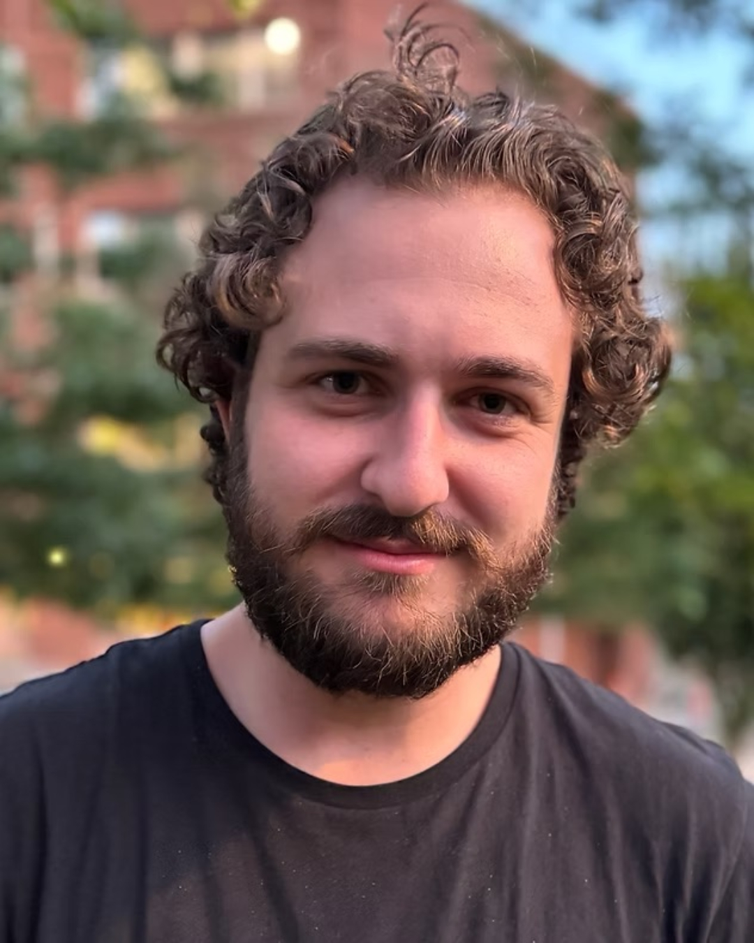 A close-up portrait of a man with curly dark brown hair styled up and to the sides, and a full dark beard. He wears a navy or black t-shirt and looks directly at the camera with a neutral expression. Behind him, a brick building and ivy-covered greenery create a soft, blurred background, suggesting an outdoor campus or institutional setting.
