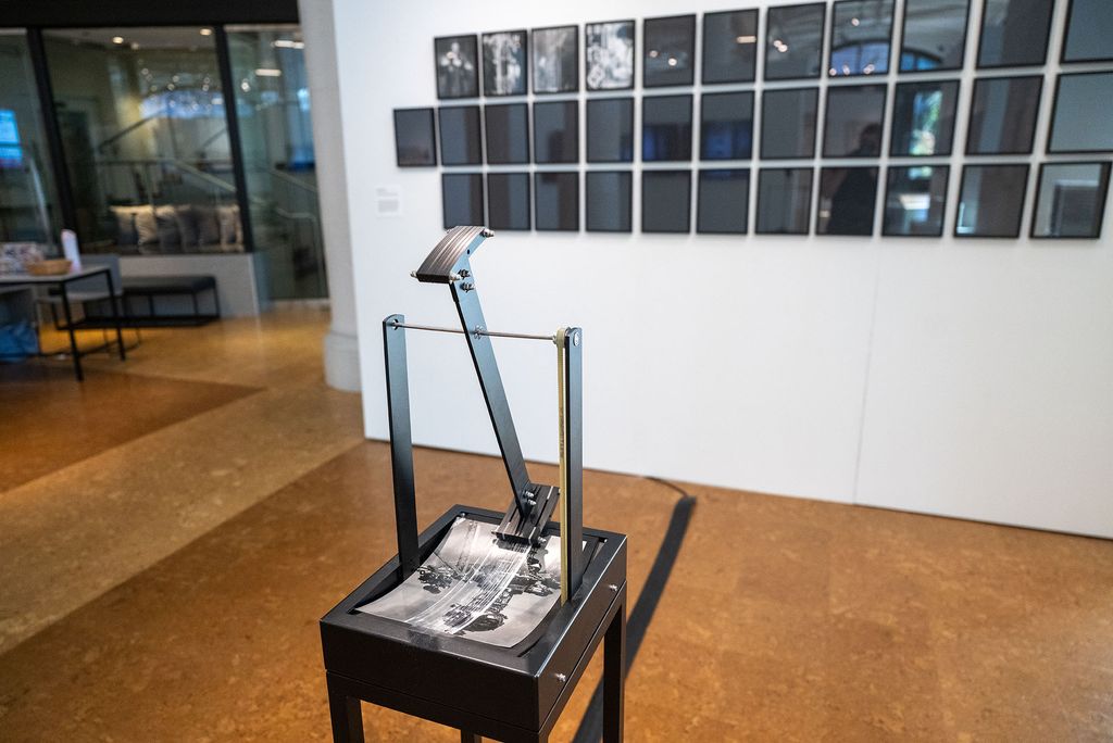 A black metal kinetic sculpture with articulated arms stands on the polished wood gallery floor, with a grid of framed photographs displayed on the white wall behind it.