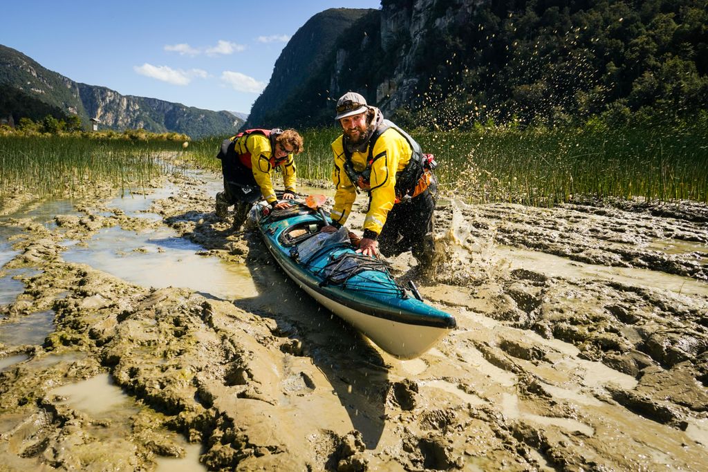 Two people wearing bright-yellow jackets and life vests, pushing a kayak through a muddy valley with grass-covered mountains on either side