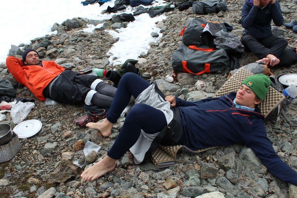 Two guys taking a nap on rocky, snow-covered ground next to backpacks and some used plates.