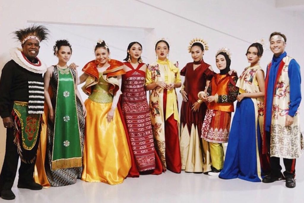Nine models are lined up, smiling and showing an array of global costumes.