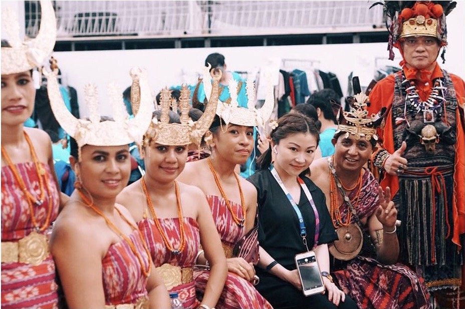 Four young women are on the left wearing wearing orange and pink strapless dresses, orange beads, a gold belt, and gold crowns. A young woman in black with an iPhone is to their right