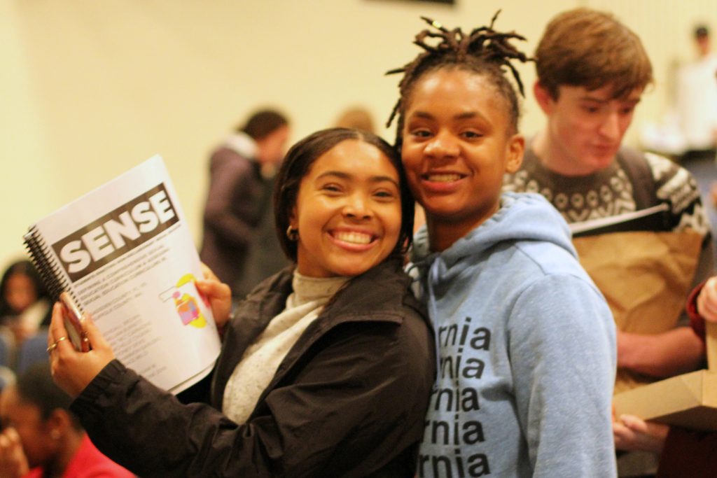 Two students looking at the camera and smiling. One is holding a bound Capstone project with the heading SENSE at the top.