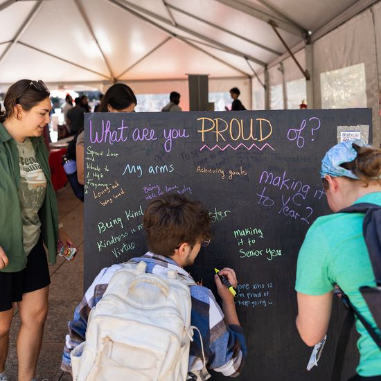 three students write on the giant chalkboard at the Religious life fair answering the question "What are you proud of?"