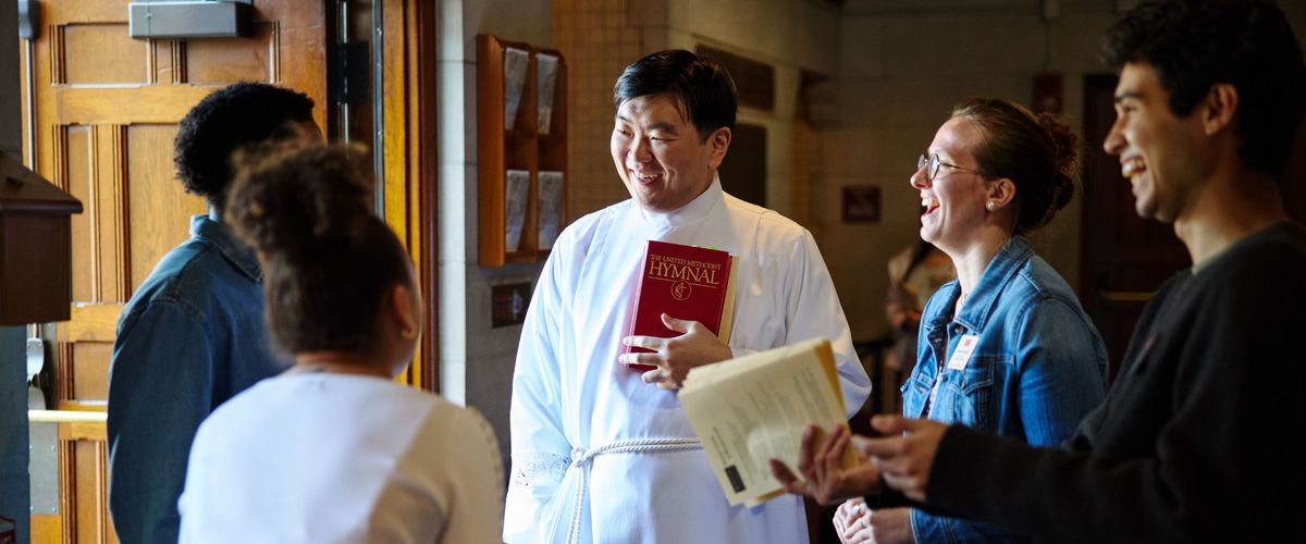 A chaplain and students stand in a circle at the chapel entrance laughing and passing out worship bulletins