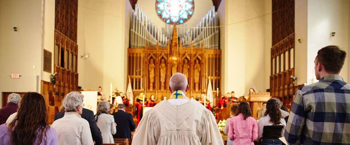 taken from the back of the sanctuary, Dean Hill walks down the aisle toward the front where the choir is singing. People of all ages sit in the pews.