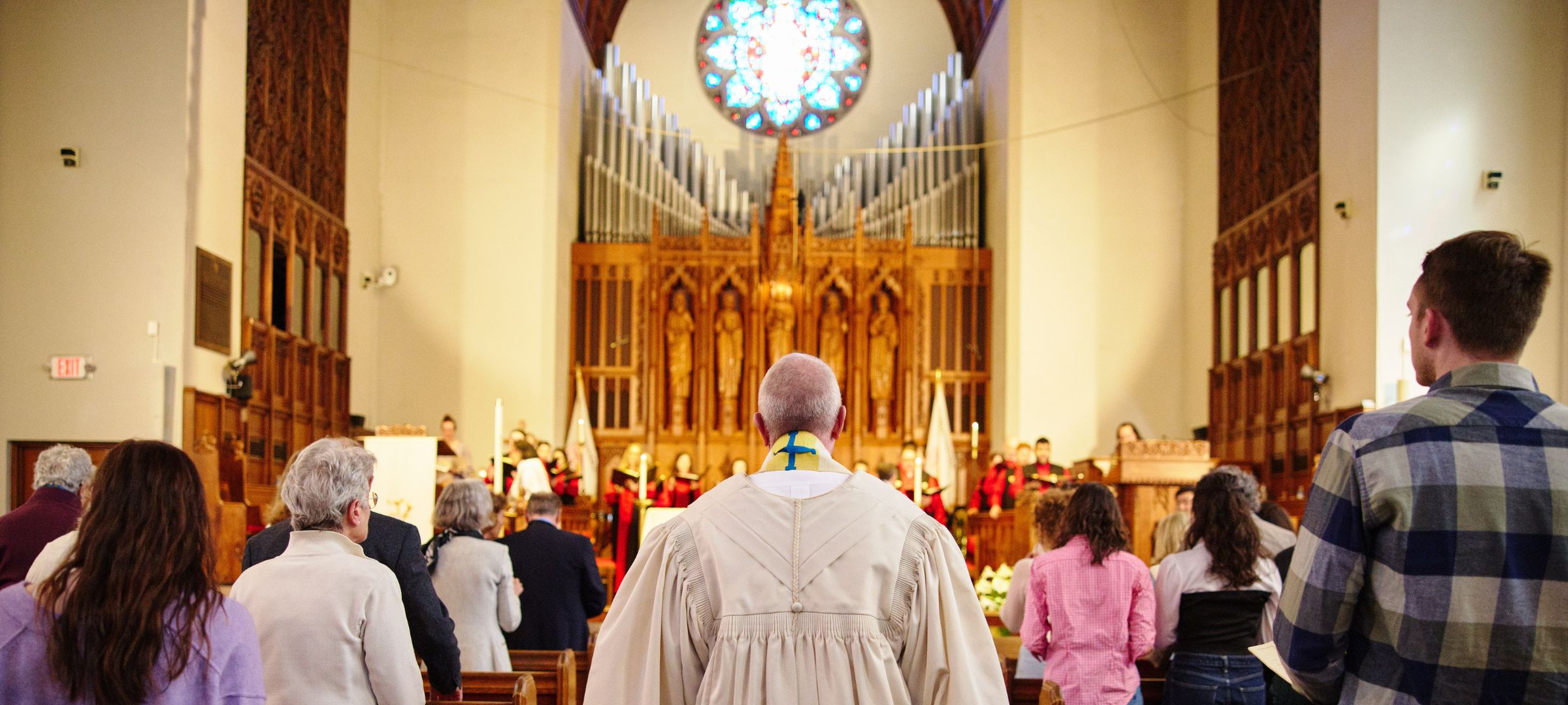taken from the back of the sanctuary, Dean Hill walks down the aisle toward the front where the choir is singing. People of all ages sit in the pews.