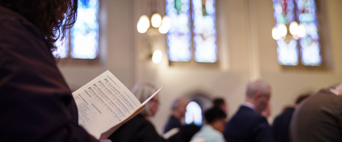 Looking over the shoulder of a congregation member. The image focuses on the words of the bulletin, while the background shows the light and colors of the stained glass. Photo intended to elicit a feeling of reflection.
